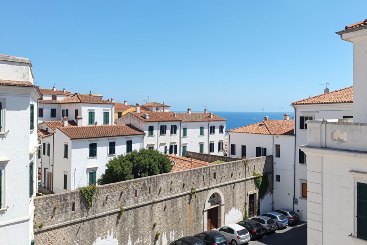 Vista panoramica del centro storico di Sperlonga con auto parcheggiate vicino alle mura bianche, cielo azzurro e scorcio del mare